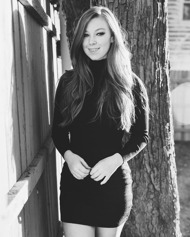 A black and white picture of Deluca in a beautiful black one-piece dress standing in front of a tree